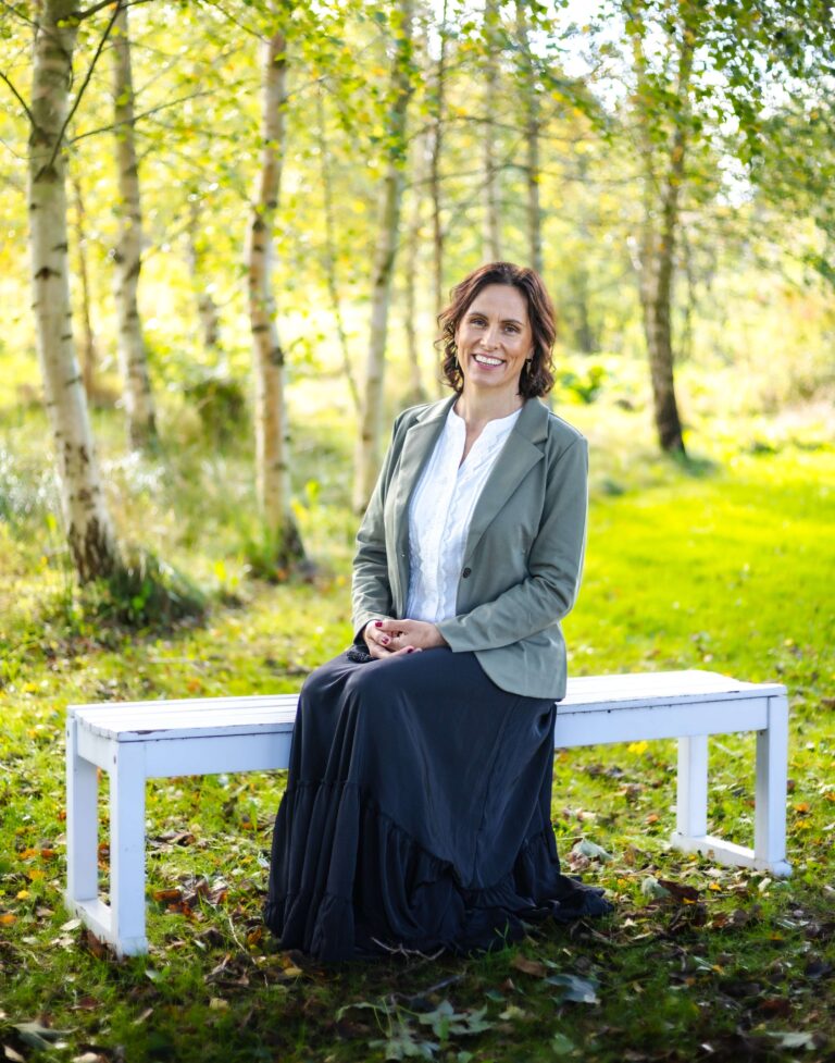 Kvinde sidder på bænk i skov, smilende ansigt.